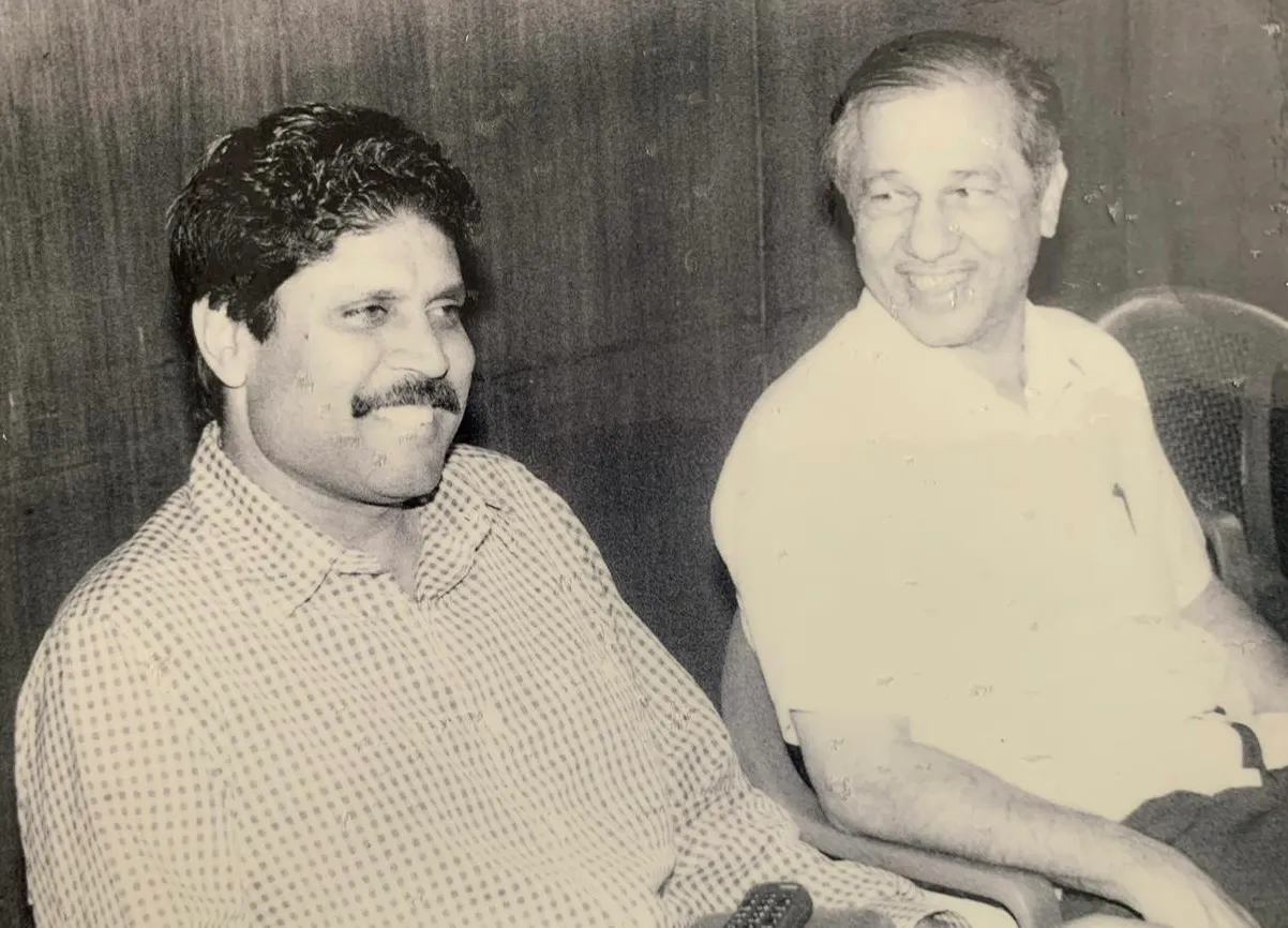 A black-and-white photograph of a smiling Kapil Dev seated beside cricket administrator Dnyaneshwar Agashe