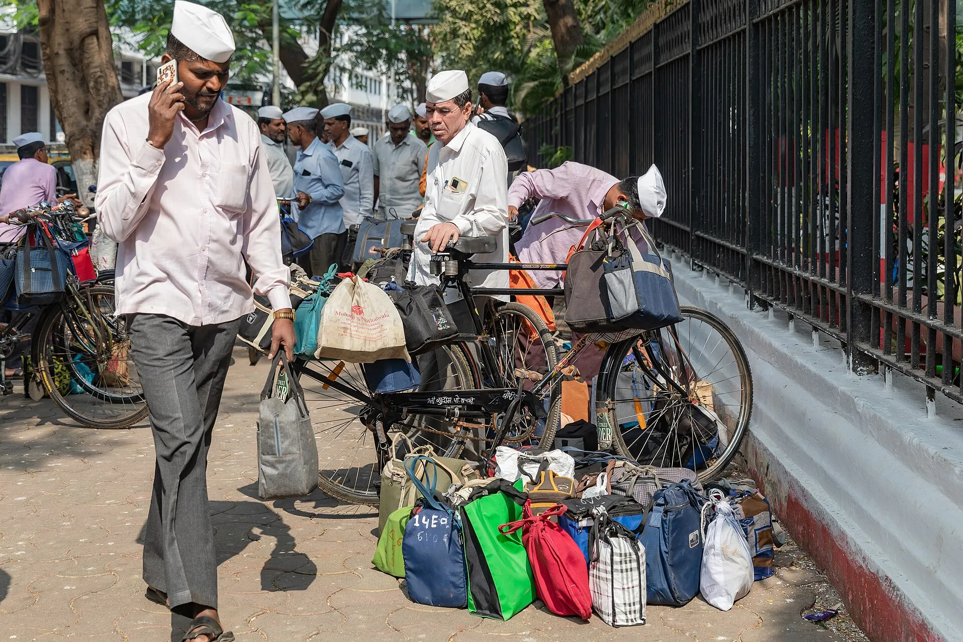 Dabbawalas in white caps on a Mumbai pavement beside bicycles hung with cloth bags of lunch boxes