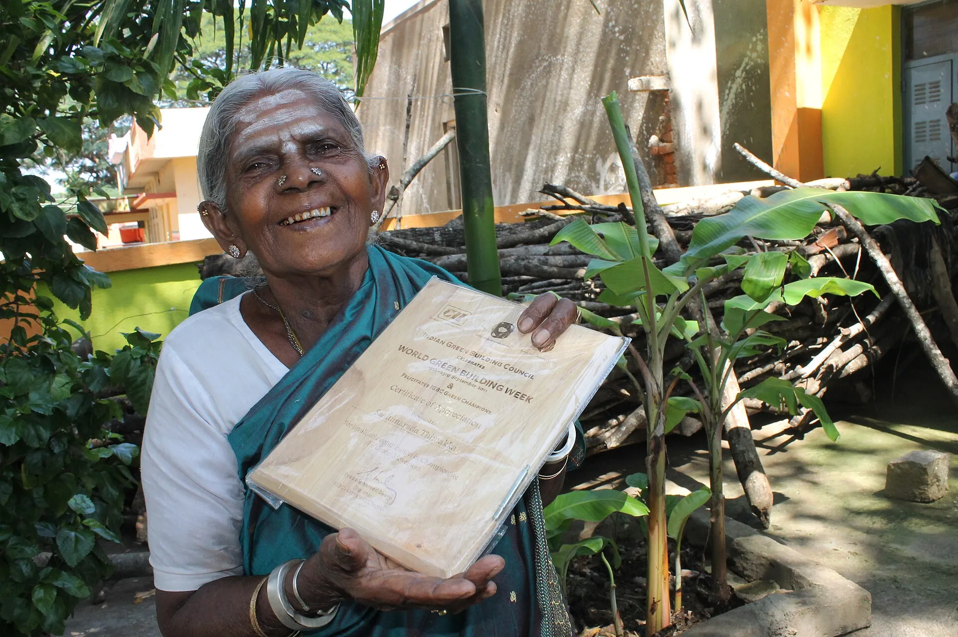 Saalumarada Thimmakka, smiling, holding up a certificate in a leafy garden